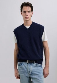 Suéter sin mangas azul marino con cuello en V, dobladillo acanalado, usado sobre una camiseta blanca, combinado con jeans de mezclilla azul claro.