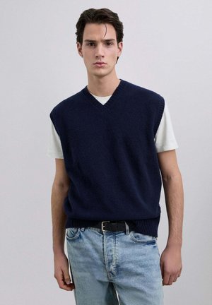 Suéter sin mangas azul marino con cuello en V, dobladillo acanalado, usado sobre una camiseta blanca, combinado con jeans de mezclilla azul claro.