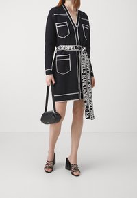 Eine Frau trägt ein schwarzes Cardigan-Kleid mit weißen Taschenumrandungen und einem Logo-Gürtel, hält eine schwarze Handtasche und trägt schwarze Riemchen-Heels.