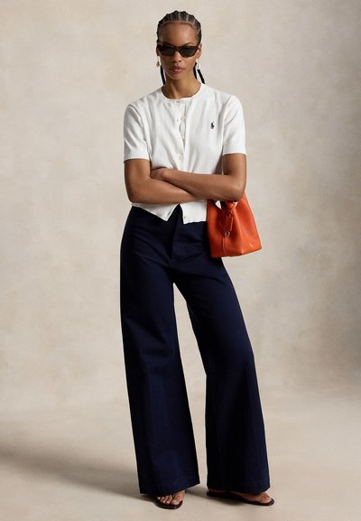 Γυναίκα που φοράει λευκό κοντομάνικο πουκάμισο με κουμπιά, σκούρο μπλε παντελόνι με φαρδύ πόδι, σκούρα γυαλιά ηλίου, κρατώντας μια πορτοκαλί τσάντα, με τα χέρια σταυρωμένα.