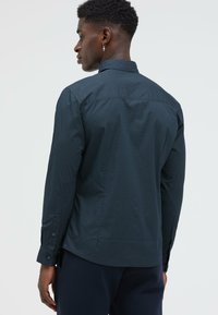 Mann, der mit dem Rücken zur Kamera steht, trägt ein dunkelblau gemustertes Langarmhemd und passende Hose, mit einem kleinen Kreuzohrring am linken Ohr.