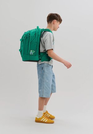 Boy wearing a large green backpack and casual clothes, standing sideways with arms slightly extended against a plain background.