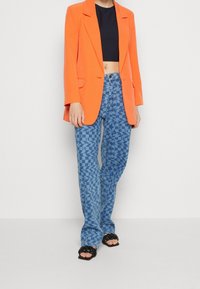 Oranžový blazer s klopovým límcem, spárovaný s modrými džíny s vzorem geometrických tvarů. Černé klouzavé sandály dotvářejí vzhled.