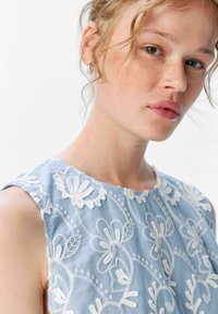 Vestido sin mangas de color azul claro con patrones florales bordados en blanco. Presenta un cuello redondo y una tela texturizada. Detalles de volantes sutiles en los hombros.