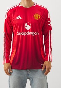 Červený dlouhý rukáv dres s bílými pruhy, s kulatým límcem, logem Manchester United a logem "Snapdragon" ve středu.