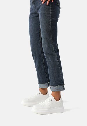Sneakers bianche con piattaforma in pelle textured, punta rotonda e design con lacci, abbinate a jeans scuri arrotolati.