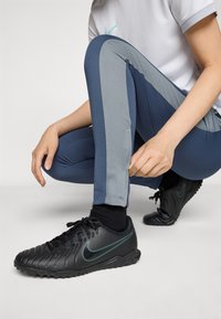 Svarte sportsko med teksturert overdel, akvamarin detaljer, kombinert med blå leggins med grå nettingpaneler, sittende posisjon, hånd som justerer stoffet.