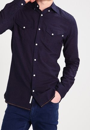 Mand iført en tætsiddende mørkeblå skjorte med hvide knapper og to brystlommer, kombineret med mørkeblå jeans.