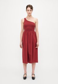 Robe rouge à une épaule avec un haut ajusté et une jupe froncée. Fabriquée en tissu lisse, longueur genoux, associée à des ballerines noires.