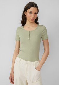 Jasnozielona bluzka z krótkim rękawem o prążkowanej strukturze z okrągłym dekoltem i pięcioma guzikami z przodu. Połączona z jasnymi spodniami.