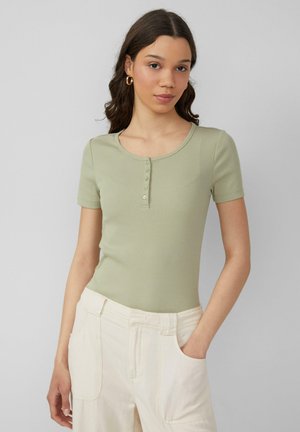 Light green ribbed short-sleeve top featuring a scoop neckline and five button accents down the front. Paired with light-colored trousers.