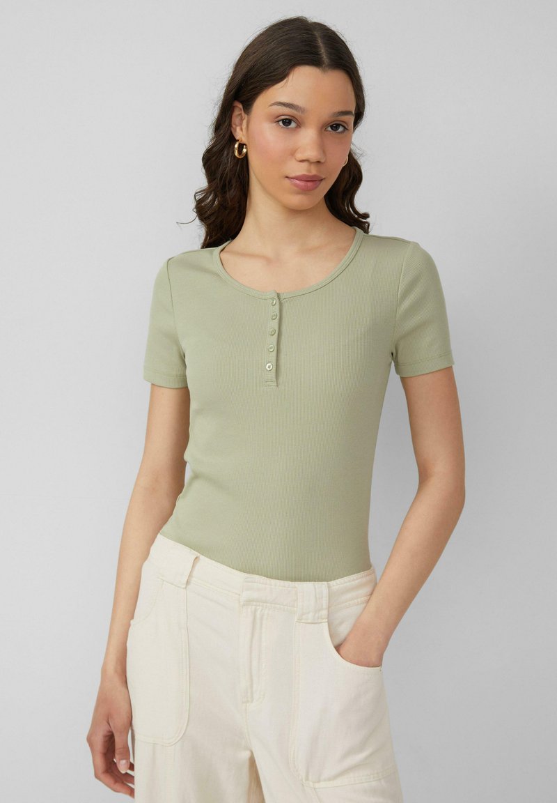 Jasnozielona bluzka z krótkim rękawem o prążkowanej strukturze z okrągłym dekoltem i pięcioma guzikami z przodu. Połączona z jasnymi spodniami.