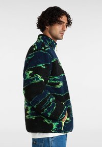 Fleecejack in zwart met groene en blauwe spiraalpatronen. Beschikt over een hoge kraag, ritszakken en een gestructureerd oppervlak.