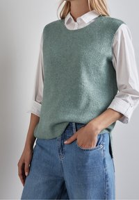 Ljusgrön stickad väst med ribbad текстur, rund halsringning, ihop med en vit skjorta med krage och blå jeans. Passformen är avslappnad med sidofickor.