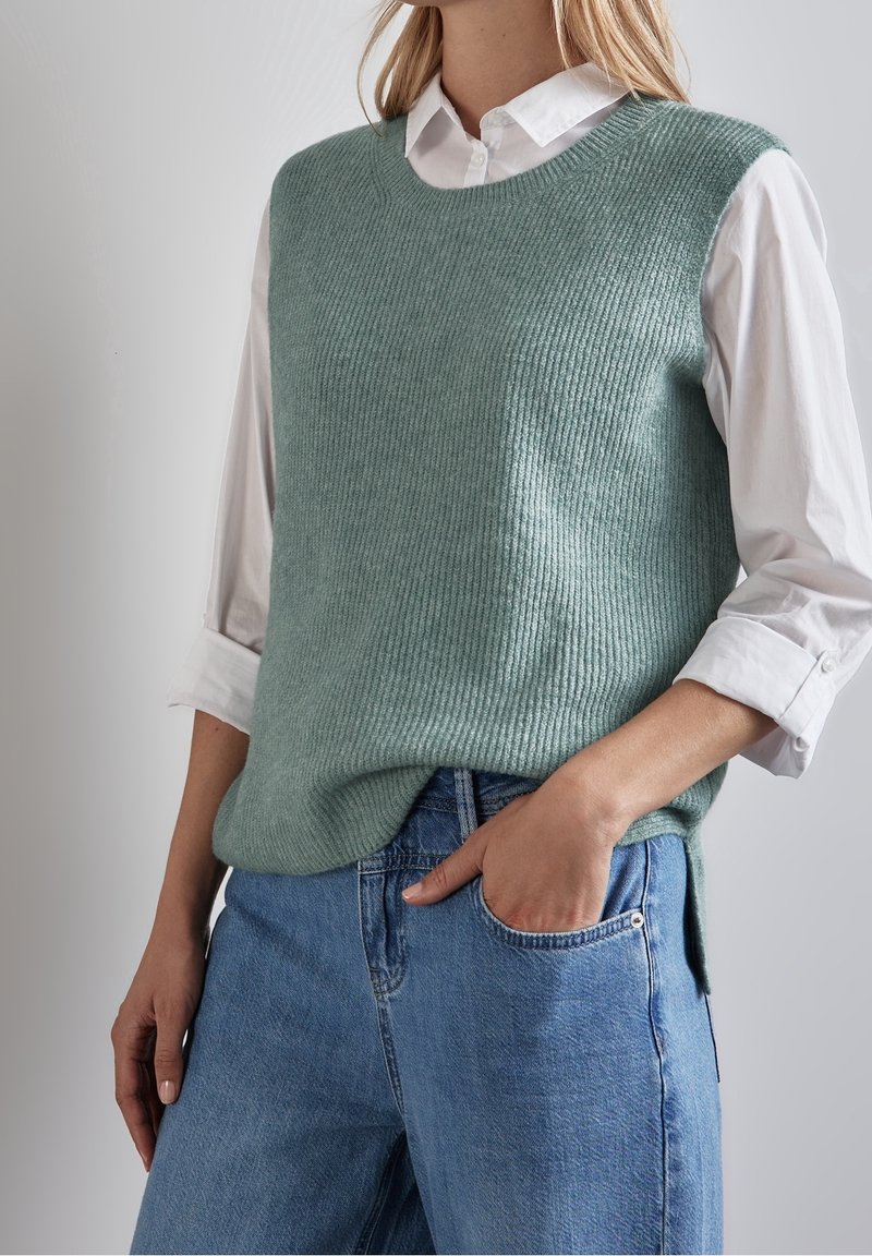 Ljusgrön stickad väst med ribbad текстur, rund halsringning, ihop med en vit skjorta med krage och blå jeans. Passformen är avslappnad med sidofickor.