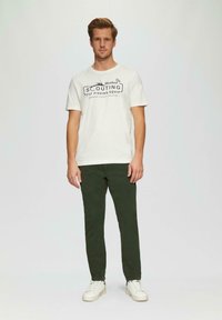 Weißes Baumwoll-T-Shirt mit schwarzem Grafiktext "Hooked On Scouting Fly Fishing Equip", kombiniert mit grünen Hosen und weißen Turnschuhen.