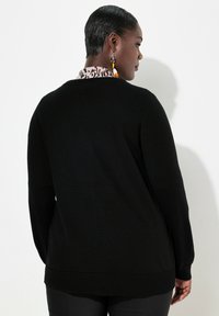 Svart stickad tröja med långa ärmar, ribbade muddkanter och nederkant, med en kontrasterande mönstrad krage som syns bakifrån.