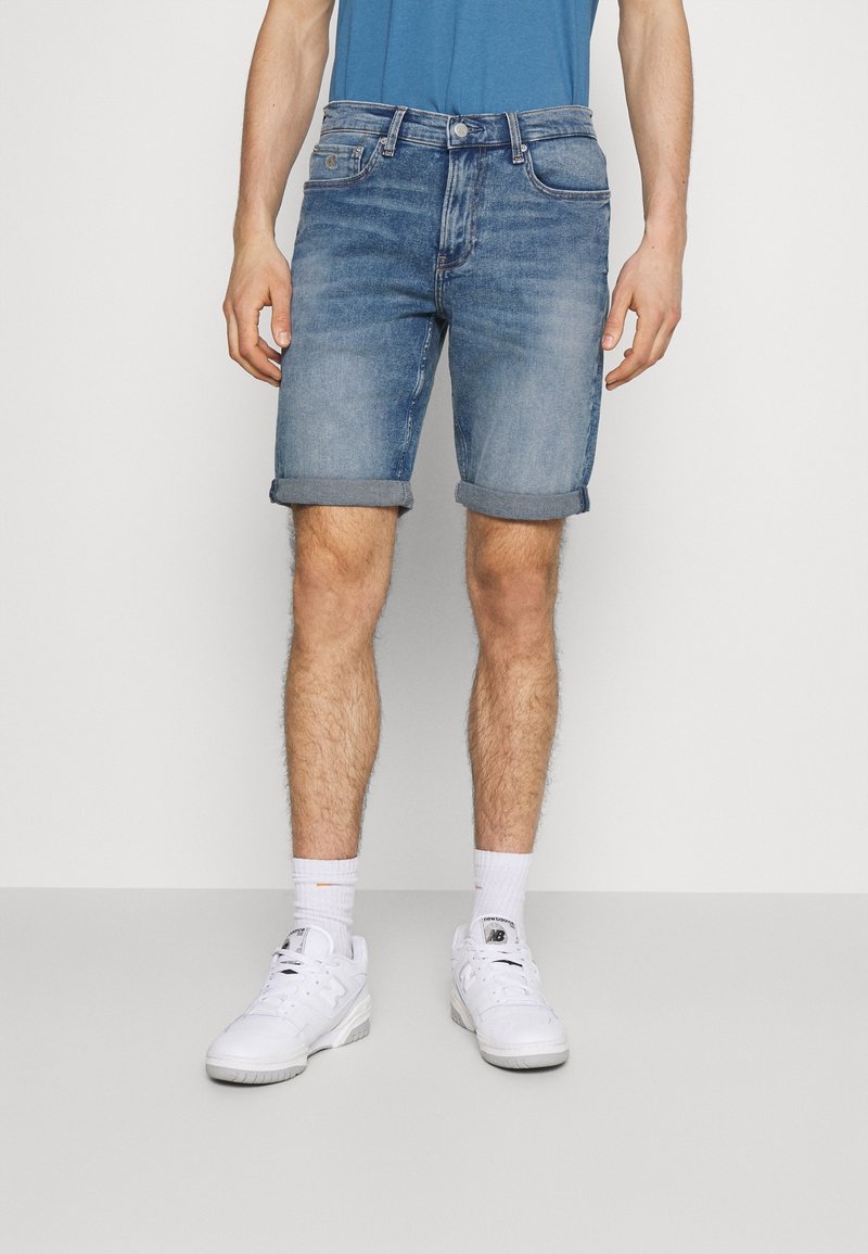 Light blue denim shorts with rolled cuffs, featuring five pockets and a button closure. Paired with white sneakers and crew socks.