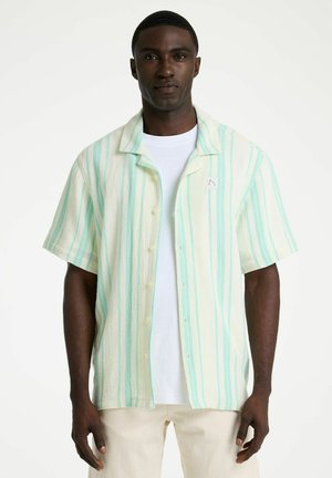 Mann trägt ein kurzärmeliges, knöpfbares Hemd mit vertikalen mintgrünen und cremefarbenen Streifen über einem weißen T-Shirt und hellen Hosen.