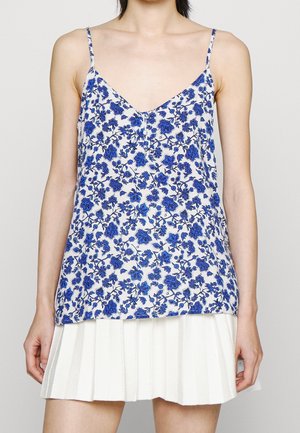 Femme portant un top blanc sans manches avec un imprimé floral bleu et une jupe plissée blanche sur un fond uni.
