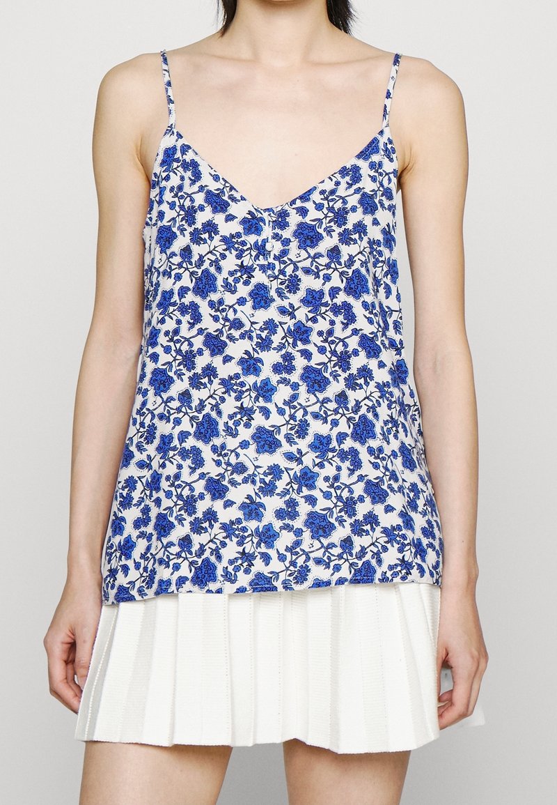 Femme portant un top blanc sans manches avec un imprimé floral bleu et une jupe plissée blanche sur un fond uni.
