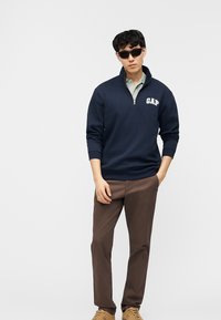 Νέος άνδρας με μαύρα γυαλιά ηλίου, μπλε σκούρο φούτερ GAP με φερμουάρ στο λαιμό, ανοιχτό πουκάμισο, καφέ παντελόνι και ταμπά παπούτσια, στέκεται με το ένα χέρι στην τσέπη.