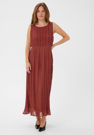 Femme portant une robe maxi plissée sans manches couleur rouille avec un col rond et des talons hauts noirs, debout avec une main sur la hanche.
