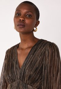 Robe rayée noire et dorée avec un décolleté en V profond, des manches longues et un léger éclat. Des boucles d'oreilles en or complètent le look.