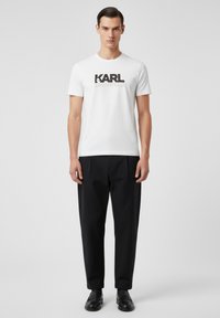 Camiseta blanca de manga corta con gráfico negro "KARL LAGERFELD", combinada con pantalones negros y mocasines negros, posando sobre un fondo neutro.