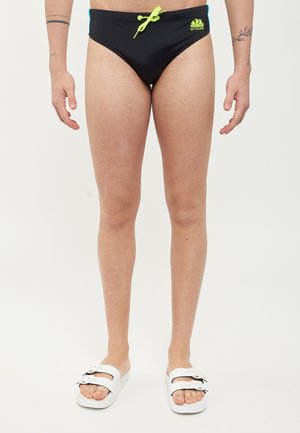 Piernas masculinas vistiendo bañador negro con cordón verde neón y logo, sandalias blancas con hebilla, de pie frente a un fondo claro liso.