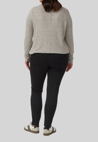 Frau in einem grauen strukturierten Langarmoberteil, schwarzen engen Hosen und weißen Sneakers mit schwarzen Streifen, die vor einem grauen Hintergrund steht.