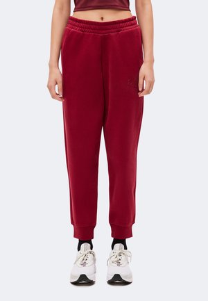 Person iført røde joggingbukser med elastisk talje og ribbet manchetter, sorte sokker, hvide sneakers og en cropped bordeaux top, stående mod en ensfarvet baggrund.
