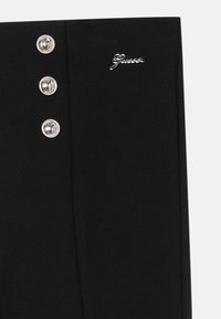 Svarta byxor med slim fit, tre silver knappdetaljer och en metallisk "Guess"-logga på midjebandet. Slät textur.