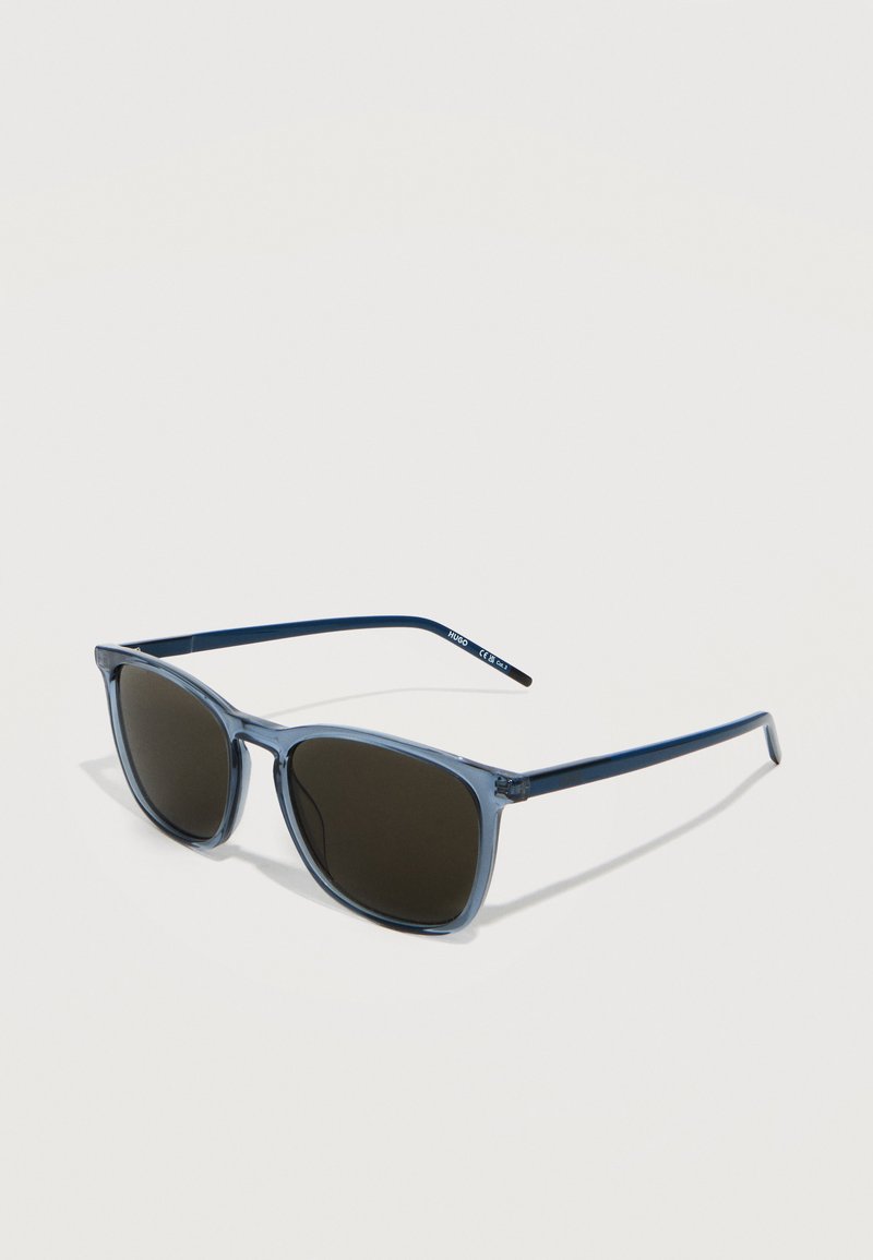 Μπλε ορθογώνια γυαλιά ηλίου με σκούρους φακούς που ακουμπούν σε λευκή επιφάνεια, με το όνομα της μάρκας "Hugo" στο πλευρό.