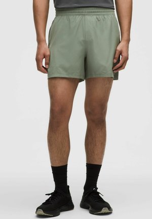 Muž oblečený v olivovo zelených športových šortkách, čiernych vysokých ponožkách a čiernych bežeckých topánkach stojaci na bielom jednoduchom pozadí.