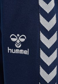 Marinblåa träningsbyxor med en vit broderad logotyp, texturerat chevronmönster på sidorna och en slät textilfinish.