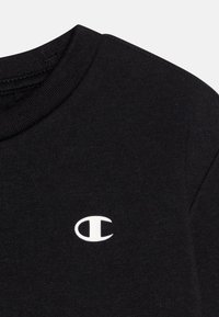 Maglietta nera in cotone con collo rotondo e un logo bianco ricamato. Texture liscia con un design aderente e uno stile minimalista.