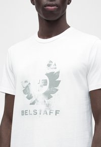 Човек, който носи бяла тениска с износено графично лого и думата "BELSTAFF" отпечатана отпред.