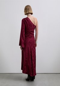 Robe bordeaux à une épaule, texture lisse avec un motif léopard discret. Silhouette longue et fluide avec une fente latérale. Bottines noires.