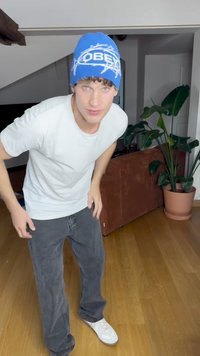Gorro azul con el logo "OBEY", camiseta blanca, pantalones vaqueros grises holgados y zapatillas blancas. Entorno interior con una planta y un sofá al fondo.