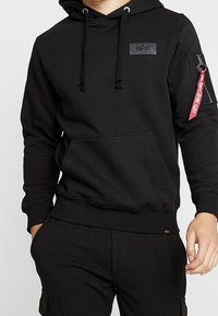 Felpa nera con cappuccio regolabile tramite coulisse, tasca a marsupio frontale e tasca con zip sulla manica dotata di una fascia rossa. Tessuto morbido e design aderente.