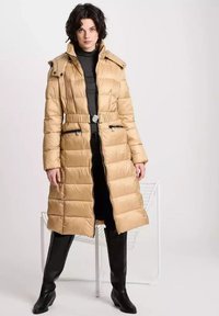Długi puchowy płaszcz w beżowym kolorze z paskiem w talii, kieszeniami na zamek błyskawiczny i wysokim kołnierzem. Tkanina ma gładką, błyszczącą teksturę.