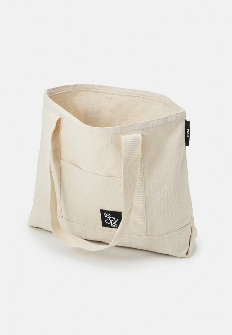 Bolso de lona en color beige con dos asas largas. Presenta un bolsillo frontal y una etiqueta negra de "reciclar" para la marca.