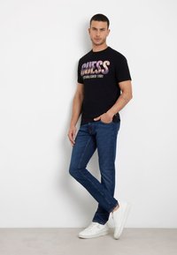 T-shirt in cotone nero con logo stampato "GUESS" in viola e oro, abbinato a jeans in denim blu e sneakers bianche, in piedi contro un muro bianco.