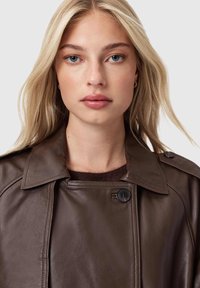 Brązowy skórzany trencz z szerokim kołnierzem, z podwójnym zapięciem na guziki i subtelnym połyskiem. Miękka faktura i strukturalny krój.