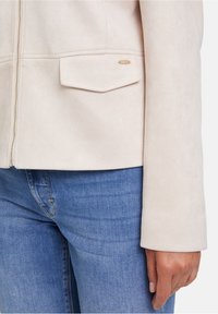 Ljust beige mockaimjäder med dragkedja, med en ficka på vänster sida, bärs över blå denimjeans. Slät textur med subtil glans.