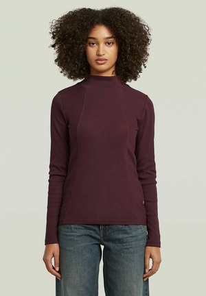 Woman with curly hair wearing a fitted, long-sleeve, dark purple top and blue jeans against a plain light background.