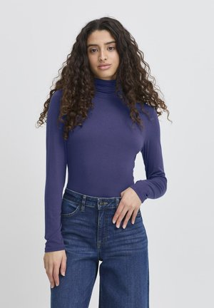 Giovane donna con lunghi capelli ricci e scuri, che indossa un dolcevita viola attillato e jeans a vita alta blu, in piedi davanti a uno sfondo semplice.