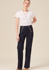 Pantalons en denim foncé taille haute avec des détails en boutons dorés, jambes droites et ourlets roulés ; associés à un t-shirt blanc uni.