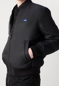 Mann in einer schwarzen Bomberjacke mit Reißverschluss, gerippten Bündchen und Saum, mit einer Hand in der Tasche und einem kleinen blauen "HUGO"-Logo auf der Brust.